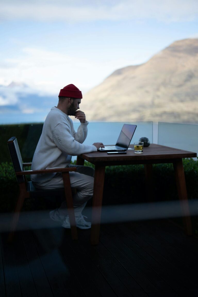 A man in casual attire works on a laptop on a terrace with stunning mountain views in Queenstown, New Zealand.