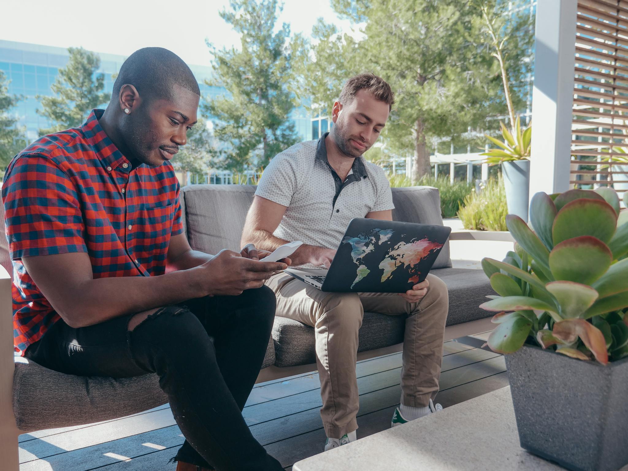 Two men having a business meeting outdoors, engaging with a smartphone and laptop, surrounded by greenery.